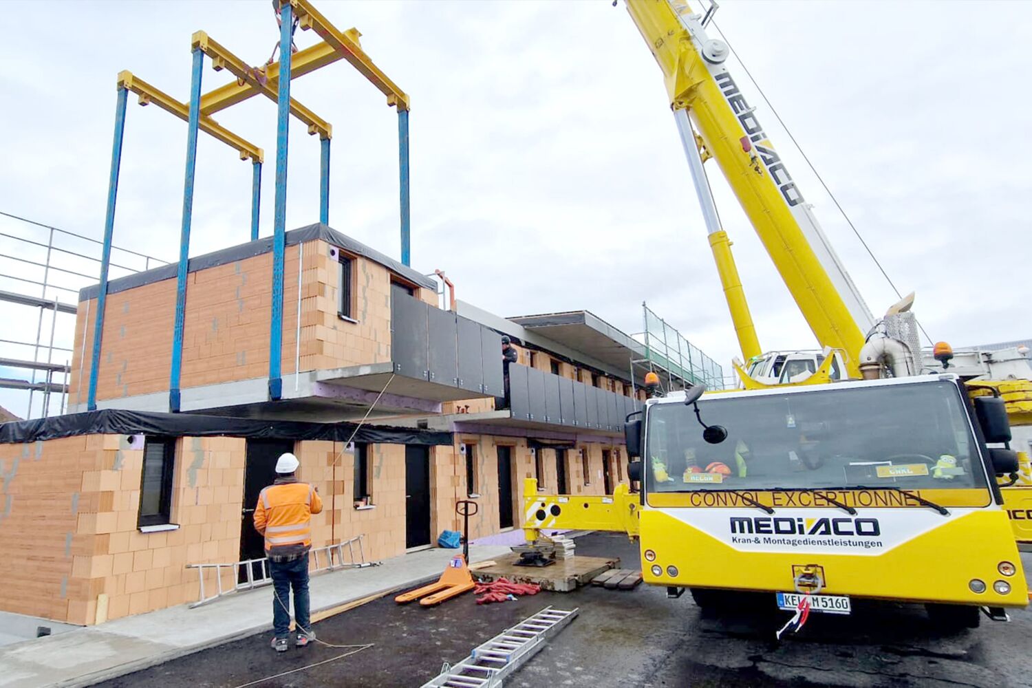 Kran hebt ein Modul auf eine Baustelle, Arbeiter sind vor Ort.